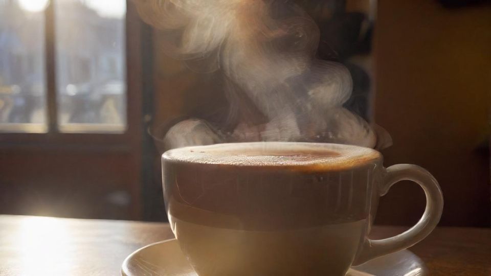 完璧に淹れられたコーヒーカップのクローズアップ、湯気が立ち上り、居心地の良い日当たりの良いカフェの雰囲気。<br  class=